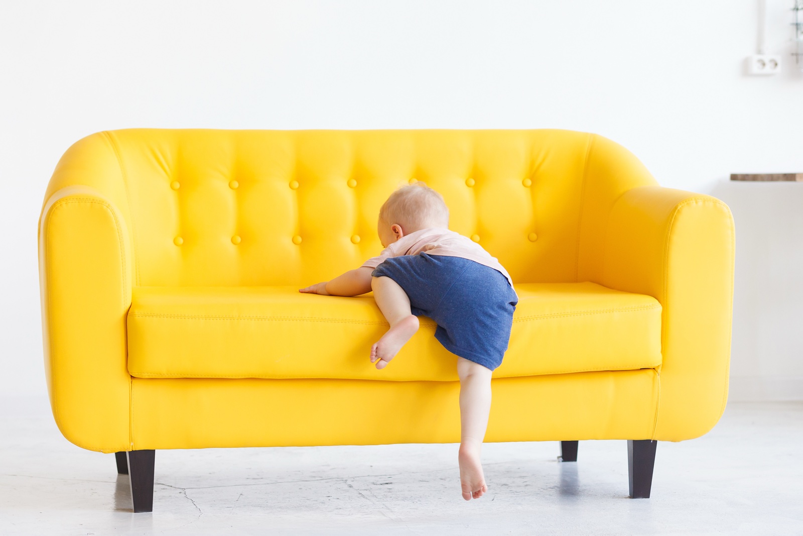 Kleinkind zieht sich an einem gelben Sofa hoch, von hinten gesehen